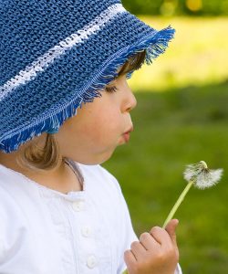 Kind mit blauer Mütze pustet eine Pusteblume